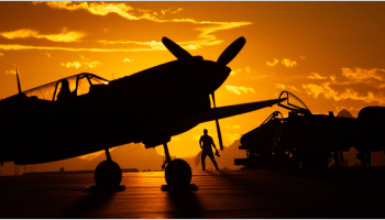 The silhouette of a military student standing near a plane can be seen against a yellow-orange sunset.