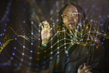 A student wearing glasses and a dark jacket holds up a hand to a glowing representation of a DNA double helix. The DNA strands curve and twist in the air in front of and around the person, giving a 3D effect.