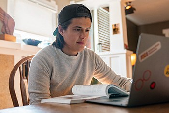 An adult student in a gray sweatshirt and backward baseball cap using a laptop at a wooden table with an open book. ASU stickers decorate the laptop.