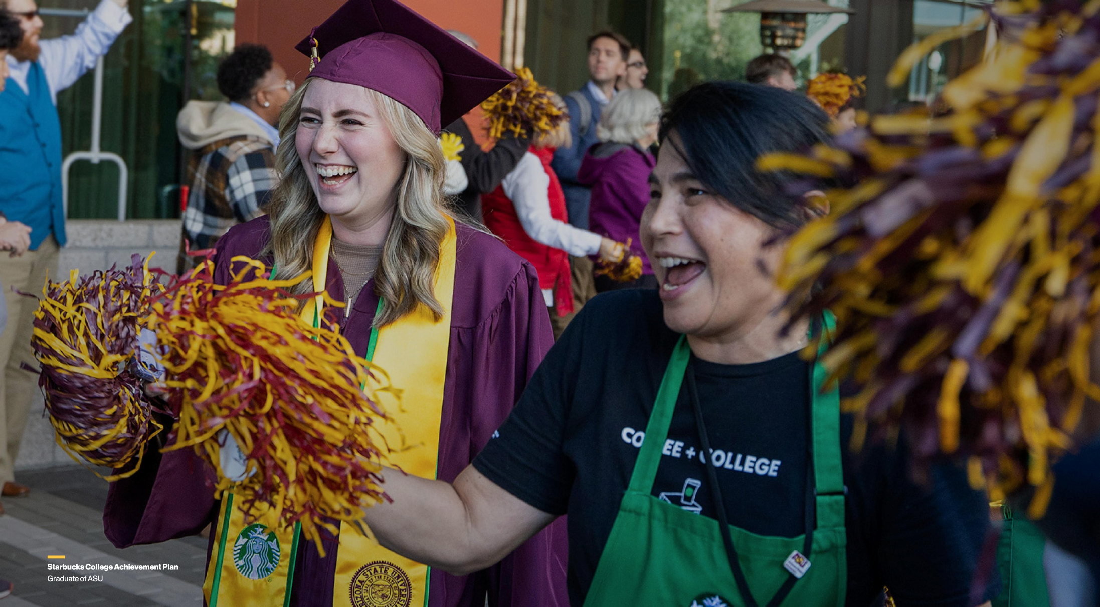 A graduate in a maroon cap and gown celebrating with pom-poms alongside another person in a green apron.