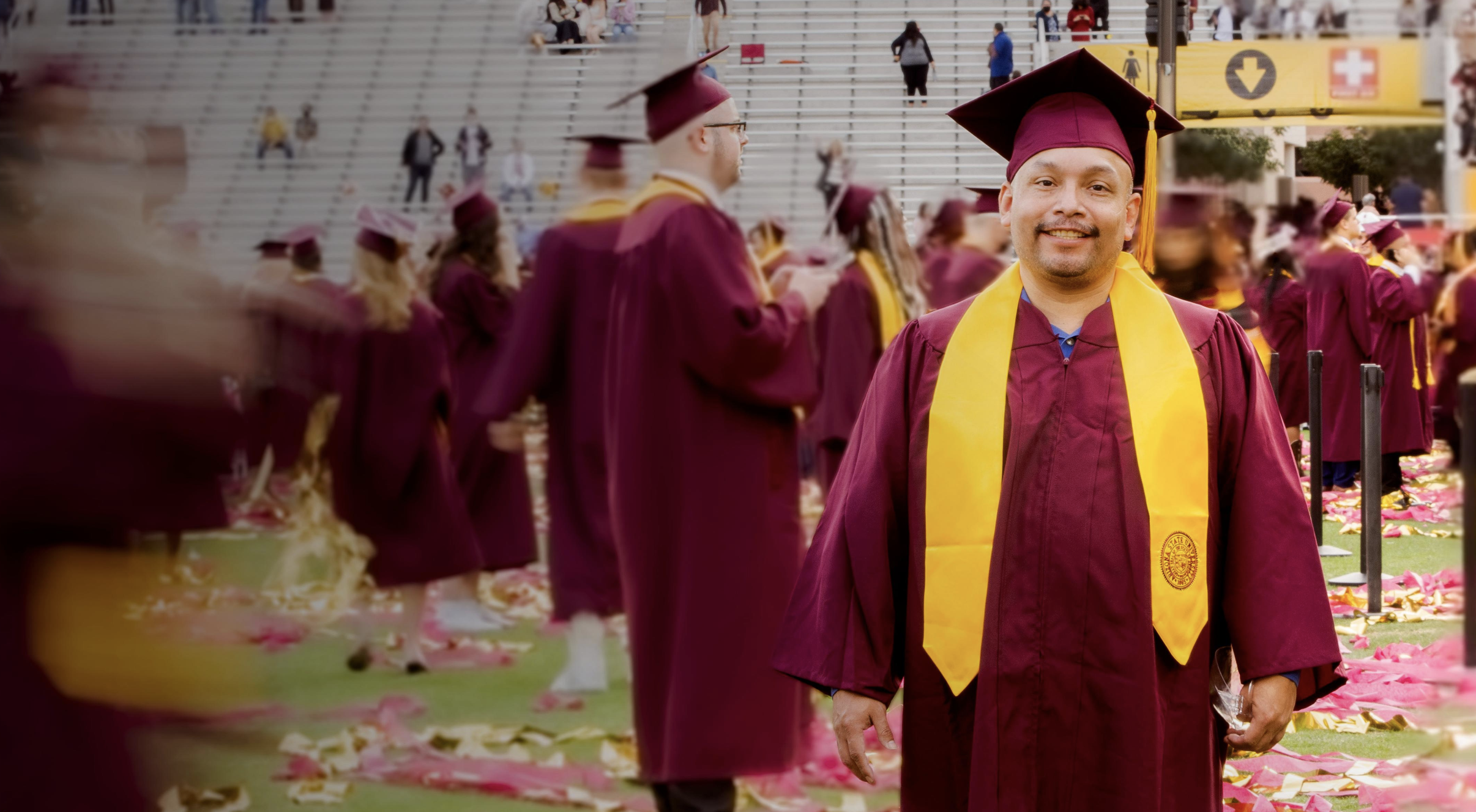 A man in a maroon cap and gown with a gold sash at a graduation ceremony, surrounded by other graduates and confetti.